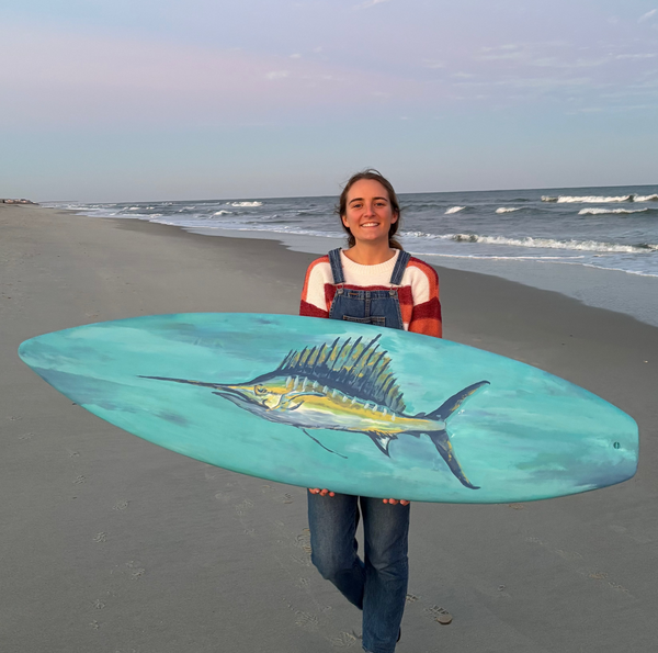 Artist holding a surfboard with a sailfish painting on surfboard in Carolina Beach